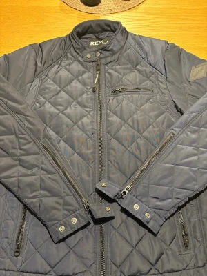 Mörkblå quiltad pufferjacka Replay - Snygg mörkblå quiltad från Replay med quiltad design och flera dragkedjor. Jackan har hög krage med tryckknappar och en bröstficka med dragkedja. Perfekt för kalla dagar och har en modern look som passar till många outfits . Ny pris 2000, pris kan diskuteras