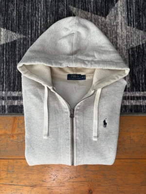Ljusgrå Polo Ralph Lauren Hoodie - Hej! 👋 Säljer en stilren ljusgrå zip-up hoodie från Polo Ralph Lauren i storlek M. Den har den klassiska svarta pony-loggan broderad på bröstet 🐎 Tröjan är 100% äkta med tvättlappar kvar. Skicket är fint, men jag vill vara helt transparent med att det finns en mycket liten prick på ena ärmen samt en liten fläck vid magfickan (se närbilder för detaljer). Det är inget som stör helhetsintrycket när den bärs! ✨ En mångsidig tröja som funkar perfekt till allt. Fraktar snabbt! 🚛📦💬💯 