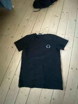 Svart t-shirt från Moncler - Svart t-shirt från Moncler med korta ärmar och rund hals. På bröstet finns Monclers ikoniska logotyp i vitt och rött. Perfekt för dig som gillar stilrena plagg med exklusiv känsla.