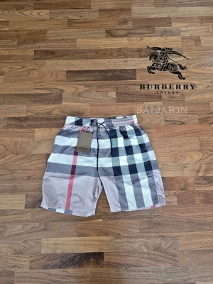 Burberry Shorts - 🩳 Burberry Shorts 🩳 Stilrena Burberry shorts med klassiskt rutmönster, perfekta för sommar, strand eller avslappnad vardag. Elastisk midja med justerbar dragsko ger bekväm passform. Tillverkade i lätt och slitstarkt material med mjuk känsla mot huden. Praktiska fickor för småsaker och Burberry-logga. Liten markering på baksidan. Shortsarna är i nyskick. 📦 Snabb leverans – Kontakta för frågor!