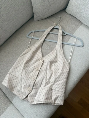 Beige randig halterneck t-shirt - Säljer en beige t-shirt med halterneck och smala svarta ränder. Modellen är ärmlös och har knappar framtill samt smock baktill för skön passform. Perfekt för dig som gillar en unik och trendig look.