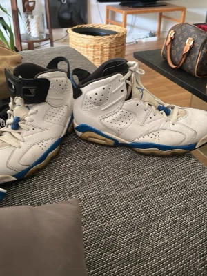 Air Jordan 6 Retro vit/blå sneakers - Säljer ett par Air Jordan 6 Retro sneakers i vitt läder med blå detaljer och perforerade paneler. Skorna har högt skaft, klassisk Jumpman-logga på plösen och en robust sula med blå och beige inslag. Perfekt för dig som gillar basketinspirerad stil.