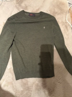 Morris merinoull - Snygg grön stickad tröja från Morris med rund hals och diskret logga broderad på bröstet. Tröjan har långärmade och en klassisk, normal passform som funkar till många olika stilar. Perfekt för dig som gillar stilrena plagg med en touch av detalj. Storlek S. Hör av er vid mera frågor.