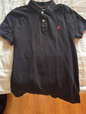 Svart Sail Racing pikétröja M - Svart pikétröja från Sail Racing med klassisk krage och knappar framtill. Diskret logga på bröstet och orange detaljer vid ärmslut och insida krage. Tröjan är tillverkad i bomull och har korta ärmar. Perfekt för dig som gillar stilrena plagg med sportig touch.