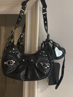 Svart axelväska med croco-mönster - Cool svart handväska med croco-mönster och silverfärgade detaljer. Väskan har flera fack, justerbar axelrem och dekorativa nitar. Medföljande småväskor och hjärtformad spegel ger extra stilpoäng. Perfekt för dig som gillar edgy och trendiga accessoarer.