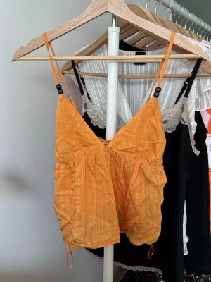 Orange linnetopp med tunna axelband - Snygg orange linnetopp med tunna axelband och träpärlor. Toppen har en lätt rynkad struktur och subtila glittriga ränder som ger extra detalj. 