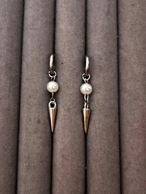 Silvriga örhängen med pärla och spike - Snygga örhängen i silverfärg med en rund vit pärla och en spetsig spike-hängande detalj. Örhängena har en enkel ring som fäste och en cool, modern design som sticker ut. Perfekt för dig som gillar att mixa klassiskt och edgy.