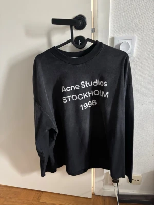 Svart hoodie Acne Studios Stockholm - Svart hoodie från Acne Studios med vit text 'Acne Studios STOCKHOLM 1996' på bröstet. Klassisk passform med rund hals och långa ärmar. Materialet ser ut att vara mjukt och bekvämt, perfekt för en avslappnad look.