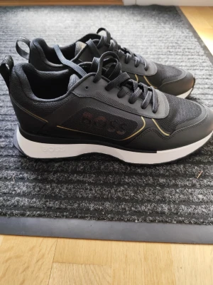 Svarta sneakers från BOSS - Stilrena svarta sneakers från BOSS med vita sulor och diskreta gula detaljer. Ovandelen är i en mix av mesh och syntetmaterial för en sportig look. Klassisk snörning och logga på sidan. Perfekta för dig som gillar en clean och modern stil.