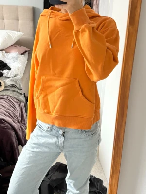 Orange cool hoodie med huva  - Säljer en snygg orange hoodie med huva och dragsnören. Tröjan har en stor magficka och ribbade muddar vid ärmslut och nederkant. Perfekt för dig som gillar en avslappnad och cool stil. Materialet känns mjukt och bekvämt, troligen bomullsblandning.