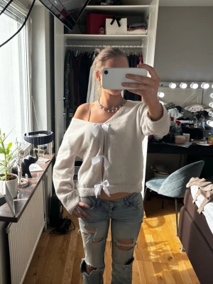 Vit stickad tröja med rosetter - Säljer en vit stickad tröja med tre dekorativa rosetter framtill. Tröjan har en avslappnad passform och är lite off-shoulder, vilket ger en cool och trendig look. Materialet är mjukt och bekvämt, perfekt för att skapa en chill outfit.