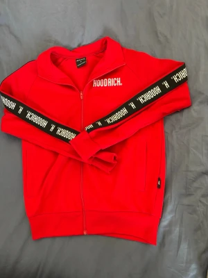 Röd ziptröja från Hoodrich - Snygg röd ziptröja från Hoodrich med vit logga på bröstet och svarta band med vit text längs ärmarna. Tröjan har hög krage, dragkedja framtill och två sidofickor. Perfekt för en streetwear-look.
