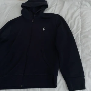 Svart hoodie från  polo  - Svart hoodie från polo  med dragkedja och klassisk vit broderad logga på bröstet. Tröjan har huva, fickor framtill och långa ärmar. Perfekt för en chill och stilren look.  Inge snören tyvärr