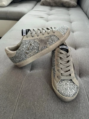 Golden Goose GGDB  - Snygga sneakers från Golden Goose med silverglitter över hela skon och beige mockadetaljer. Klassisk låg modell med GGDB-logga på tungan och stjärna på sidan. Perfekt för dig som vill sticka ut med ett par riktigt coola skor.
