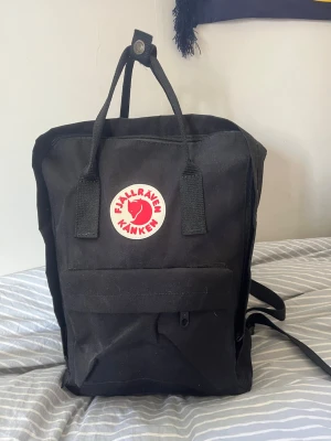 Svart Kånken ryggsäck från Fjällräven - Klassisk svart Kånken ryggsäck från Fjällräven med det ikoniska runda röda och vita logotypmärket framtill. Ryggsäcken har två topphandtag, justerbara axelband, en stor huvudficka med dragkedja och en mindre framficka. Tillverkad i slitstarkt syntetmaterial.