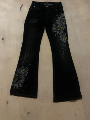 Svarta utsvängda jeans med brodyr - Unika svarta jeans med utsvängda ben och stora broderade blommor i vitt, grått och grönt på ena benet. Femficksmodell med knapp och dragkedja. Perfekta för dig som vill sticka ut med coola detaljer och retrovibbar. Står ingen storlek i byxorna men skulle gissa på S