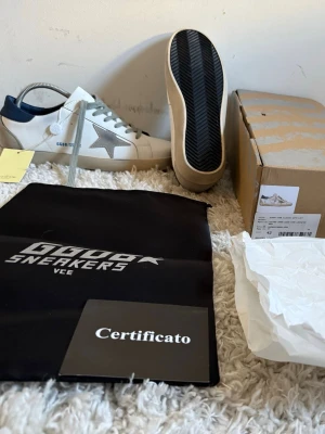 Golden Goose vita sneakers med stjärna - Golden Goose sneakers i vitt läder med grå mockadetaljer och ikonisk stjärna på sidan. Skon har mörkblå hälkappa, beige sula och grå skosnören. Klassisk låg modell med cool vintagekänsla och märkets logga på sidan.