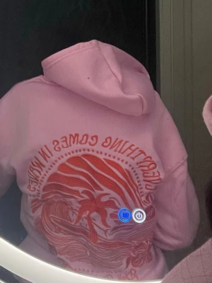 Rosa hoodie med tryck och ficka - Säljer en ljusrosa hoodie med stor ficka framtill och ett coolt rött tryck på ryggen med texten 'Everything comes in waves' och vågmotiv. Hoodien har luva och är i mjukt material, perfekt för en avslappnad stil.storlek M 