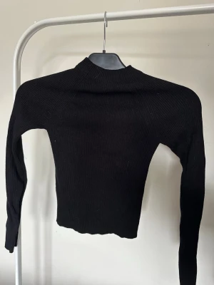 Svart ribbstickad topp med cut-out - Trendig svart ribbstickad topp med lång ärm och cut-out detalj framtill. Toppen har en hög hals och knytband vid bröstet för en cool och unik look. Materialet är stretchigt och sitter tajt mot kroppen. Perfekt för dig som vill ha en edgy stil.