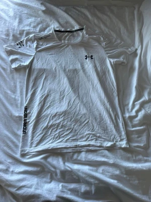 Vit Under Armour tränings-tshirt - Säljer en vit tränings-tshirt från Under Armour med svart logga på bröstet och tryck längs sidan. Materialet är lätt och andas, perfekt för löpning eller gym. Rund hals och korta ärmar, tillverkad i syntetiskt funktionsmaterial.