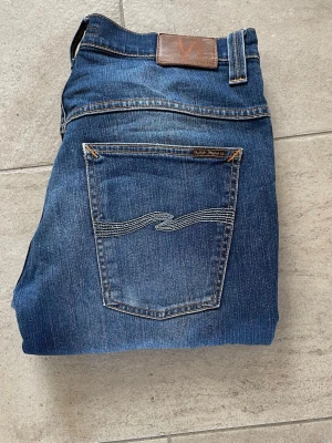 Blå jeans nudie  - Snygga blå jeans från nudie jeans med klassisk femficksdesign och coola broderade bakfickor. Jeansen har en normal passform och är gjorda i mjukt denim-material. Perfekta för dig som vill ha ett stilrent och tidlöst plagg.