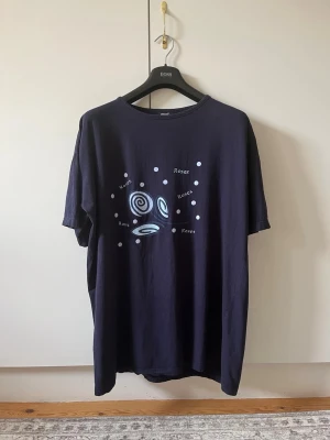 Mörkblå t-shirt med tryck från Gazzi strl XL (46/48) - Mörkblå t-shirt från Gazzi i 100% bomull. Framsidan har ett tryck med spiraler, prickar och texten 'Roses' i vitt och ljusblått. Klassisk passform med rund hals och korta ärmar. Storlek XL (46/48).