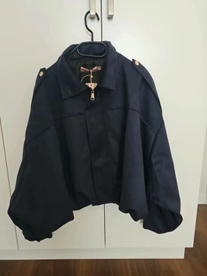 Oversize mörkblå jacka med guldzip XL  - Cool oversize jacka i mörkblått med breda ärmar och axelknappar. Jackan har en guldig dragkedja framtill och klassisk krage. Materialet ser ut att vara kraftigt tyg, perfekt för att skapa en trendig look.