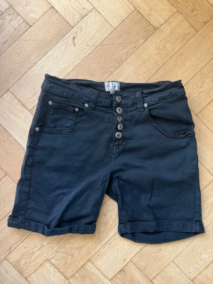 Svarta shorts med knappar - Svarta shorts med fem knappar framtill, uppvikta benslut och klassiska fickor både fram och bak. Mycket stretchiga!