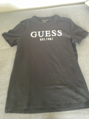 Svart t-shirt från Guess med tryck - Svart t-shirt från Guess med vit logotyp och text 'EST. 1981' på bröstet. Modellen har rund halsringning och korta ärmar. Slim fit-passform och tillverkad i mjuk bomull som är skön att bära.