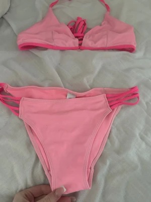 Rosa bikini med knyt - Snygg rosa bikini med trekantsöverdel och knyt i nacken och ryggen. Underdelen har smala band i sidorna för en trendig look. Perfekt för stranden eller poolen, och färgen poppar verkligen i solen.