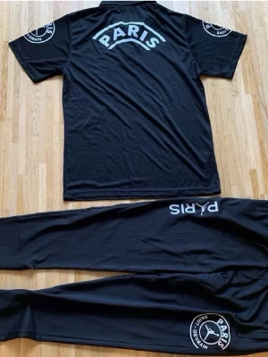 Svart PSG x Jordan träningsset - Säljer ett svart träningsset från Jordan x Paris Saint-Germain med tydliga vita loggor på både tröja och byxor. Setet består av en kortärmad tröja och matchande byxor i mjukt funktionsmaterial. Perfekt för dig som gillar sportig stil och vill representera PSG.