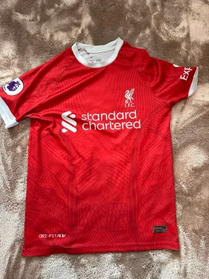 Liverpool FC röd matchtröja Nike - Säljer en röd Liverpool FC matchtröja från Nike med vita detaljer vid hals och ärmslut. Tröjan har klubbens logga, sponsortryck och Premier League-märke på ärmen. Materialet är Dri-FIT för att hålla dig torr under träning eller match.