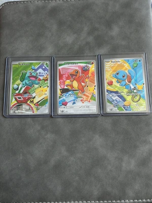 Pokémonkort Bulbasaur Charmander Squirtle - Säljer tre Pokémonkort med Bulbasaur, Charmander och Squirtle. Korten har färgglada illustrationer och ligger i skyddande plastfickor. Perfekt för samlare eller Pokémonfans som vill utöka sin samling med klassiska starter-Pokémon.