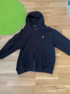 Svart hoodie från Polo Ralph Lauren - Säljer en svart hoodie med dragkedja från Polo Ralph Lauren. Hoodien har huva, två fickor framtill och den klassiska vita logon broderad på bröstet. Perfekt för dig som gillar stilrena och bekväma plagg.