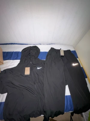 Nike Miller - Säljer ett svart träningsset från Nike med jacka och byxor. Jackan har huva, dragkedja och vit Nike-logga på bröstet. Byxorna har resår i midjan och vit logga på benet. Perfekt för sport och träning, tillverkad i lätt och funktionellt material.