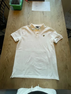 Vit polo t-shirt från Lyle & Scott - Vit polo t-shirt från Lyle & Scott med klassisk krage och två knappar framtill. Storleken är för 15-16 år. Den är aldrig använd så den är i ett helt nytt skick.Tveka inte om du har frågor!