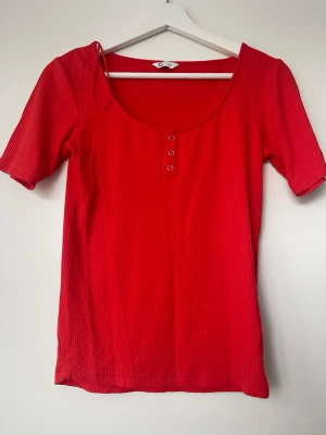 Ribbad topp med knappar - Trendig röd ribbad topp med korta ärmar och rundad urringning. Toppen har tre dekorativa metallknappar framtill och är tillverkad i ett mjukt, stretchigt material som sitter snyggt på kroppen. Perfekt att styla med jeans eller kjol.