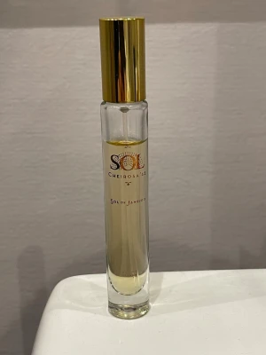 Sol de Janeiro Cheirosa 62 parfym - Sol de Janeiro Cheirosa 62 är en parfym i en smal, cylindrisk glasflaska med guldfärgat lock. Flaskan är transparent och visar den ljusgula vätskan inuti. Märkets logga och namn är tryckt i guld och svart på flaskan. 