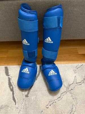 Blå benskydd och fotsskydd Adidas - Säljer ett par blå benskydd och fotsskydd från Adidas, perfekta för kampsport som karate eller taekwondo. De har justerbara kardborreband för optimal passform och är tillverkade i slitstarkt syntetmaterial. WKF-godkända och med tydlig Adidas-logga på framsidan.