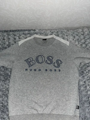 Grå Hugo Boss hoodie med logga - Snygg grå hoodie från Hugo Boss med stor BOSS-logga i blått på bröstet och vita detaljer vid axlarna. Tröjan har ribbade muddar och är tillverkad i mjukt material som känns skönt mot huden. Perfekt för dig som gillar stilrena och trendiga plagg.PRISET KAN DISKUTERAS 