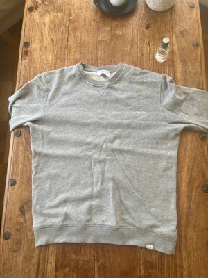 Grå sweatshirt från Les Deux - Säljer en stilren grå sweatshirt från Les Deux med broderad logga på bröstet. Tröjan har rund hals, ribbade muddar och är gjord i mjukt material som är perfekt för en avslappnad look. 