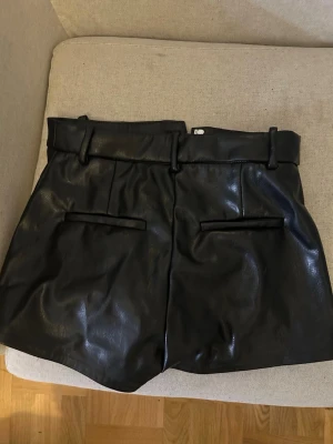 Svarta läder shorts  - Snygga svarta läder shorts med hög midja. De har bälteshällor, två fickor fram och en dragkedja. Perfekta för dig som vill ha en edgy och trendig stil. Passformen är normal och materialet är syntet med en glansig finish.