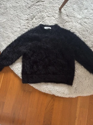 Svart fluffig stickad tröja från Zara - En svart stickad tröja från Zara med fluffig och mjuk textur. Tröjan har rund halsringning och långa ärmar, perfekt för att hålla sig varm och se stylish ut. Materialet är syntetiskt och ger en cool, trendig look.