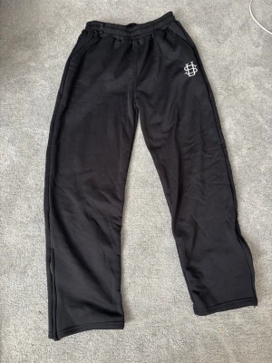 Svarta mjukisbyxor med logga - Svarta mjukisbyxor från SikSilk med vit broderad logga på vänster höft. Byxorna har elastisk midja och raka ben, tillverkade i mjukt material som är perfekt för chill och avslappnade dagar.
