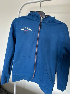 Blå zip-hoodie från Sebago - Blå hoodie med dragkedja från Sebago, med tryck 'Sebago 1946' på bröstet. Klassisk huva och känguruficka framtill. Mjuk bomullskänsla och ribbade muddar vid ärmslut och nederkant. Perfekt för en avslappnad och sportig stil.
