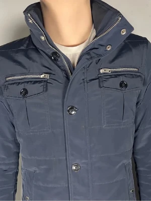 J.Lindeberg Bailey Field jacket - Snygg mörkblå pufferjacka från J.lindeberg som har en hög krage, knappar och dragkedjor på bröstfickorna i snygg silver finish . Jackan har ett quiltat mönster och en liten logotyp på ärmen. Perfekt för kalla vinterdagar. Jackan har en modern, stilren look. Skriv vid fler funderingar😄