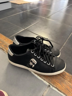 Svarta Karl Lagerfeld sneakers - Snygga svarta sneakers från Karl Lagerfeld med silverdetaljer på hälen och en ikonisk Karl Lagerfeld-logga på sidan. Skorna har svarta skosnören, vita sulor och är tillverkade i läder. Perfekta för dig som vill ha en stilren och trendig look.