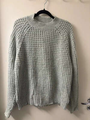 Ljusgrön stickad tröja med struktur - Säljer en ljusgrå stickad tröja med grovt strukturerat mönster och ribbade muddar vid hals, ärmslut och nederkant. Tröjan har en rund halsringning och långa ärmar, perfekt för att hålla sig varm under kyliga dagar.