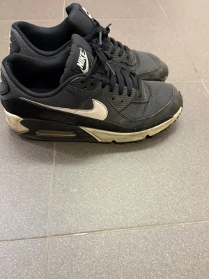 Nike Air Max 90 - Säljer ett par klassiska Nike Air Max sneakers i svart med vita detaljer och logga. Skorna har en låg profil, mesh och syntetmaterial på ovansidan samt en synlig Air Max-dämpning i sulan. Perfekt för dig som gillar sportig och stilren design.