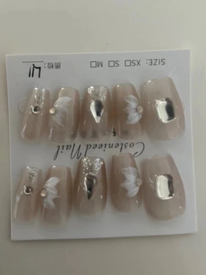 Beige press-on naglar med glitter - Snygga press-on naglar i beige med silverglitter, vita blommor och metallicdetaljer. Några naglar har små pärlor för extra bling. Perfekt för dig som vill ha en trendig och elegant look. Kommer i flera storlekar för bästa passform.